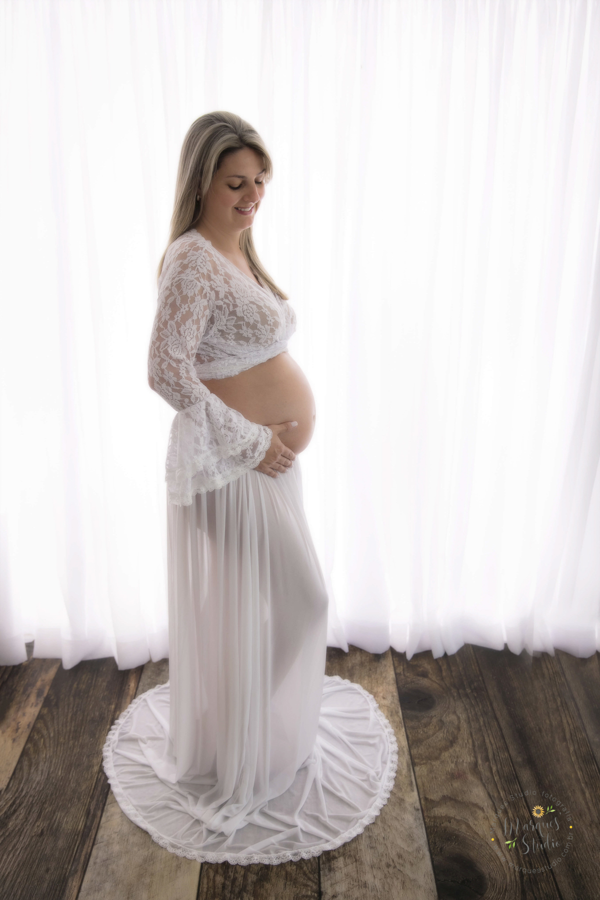 Fotografia tirada em studio localizado na Pompeia - São Paulo, SP. Na foto a futura mamãe está com usando um lindo conjunto de saia e top branco, ela está de perfil em frente a uma linda cortina branca, segurando a sua barriga