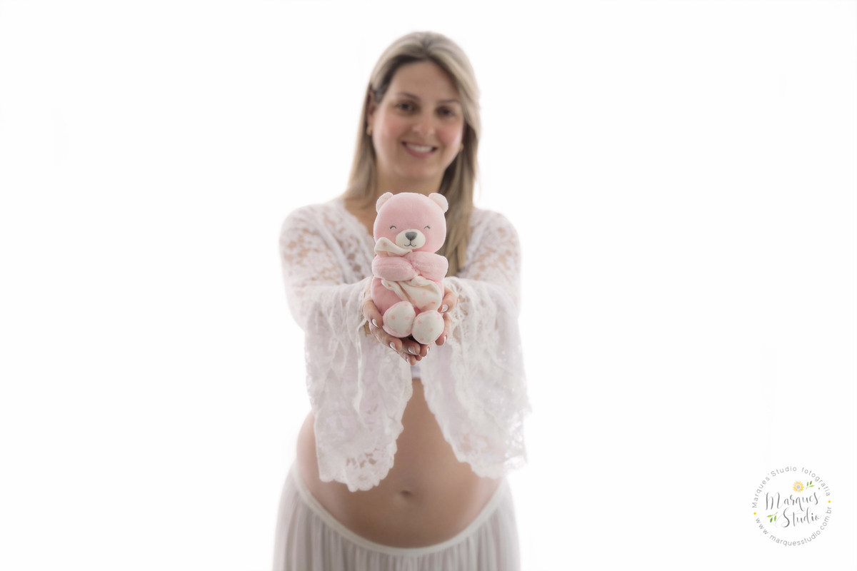 Fotografia tirada em studio localizado no Morumbi - São Paulo, SP. Na foto a futura mamãe está com usando um lindo conjunto de saia e top branco, em frente a uma linda cortina branca, segurando um urso de pelúcia rosa. 