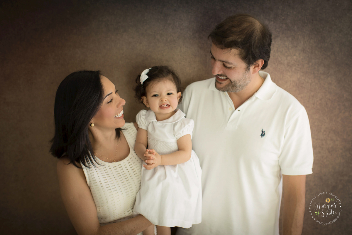 Fotografia em familia realizada no ensaio smash the cake da Lais. Na foto temos a Lais no colo da mamãe e do papai. Ela está rindo muito e os papais estão rindo olhando pra ela.