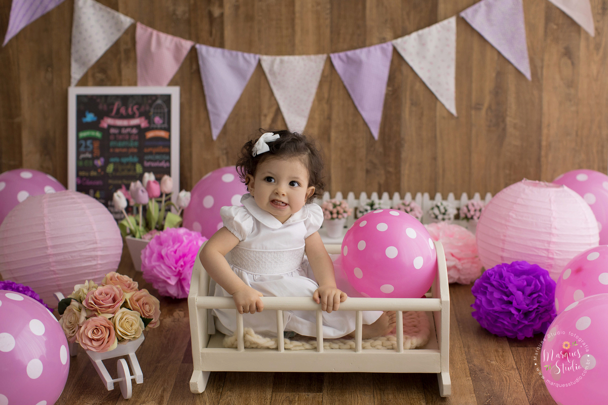 Fotografia registrado em ensaio smash the cake em studio localizado na Pompeia - São Paulo, SP. Na foto temos a Lais sentada em um berço bege, ela usa um vestido e um lacinho branco. No fundo um cenário rosa cheio de balões, flores, bandeirolas e quadros 