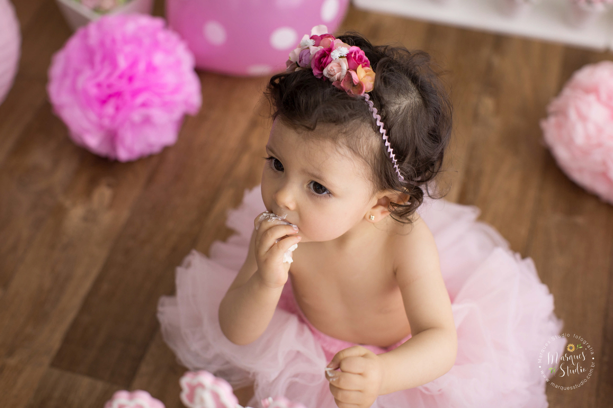 Fotografia registrada em studio localizado em Osasco - São Paulo, SP. Na foto temos a Lais sentada no chão, usando uma saia de tule rosa, com as mãos e o rosto cheios de bolo branco. Ela está comendo  mais bolo com a mão na boca.