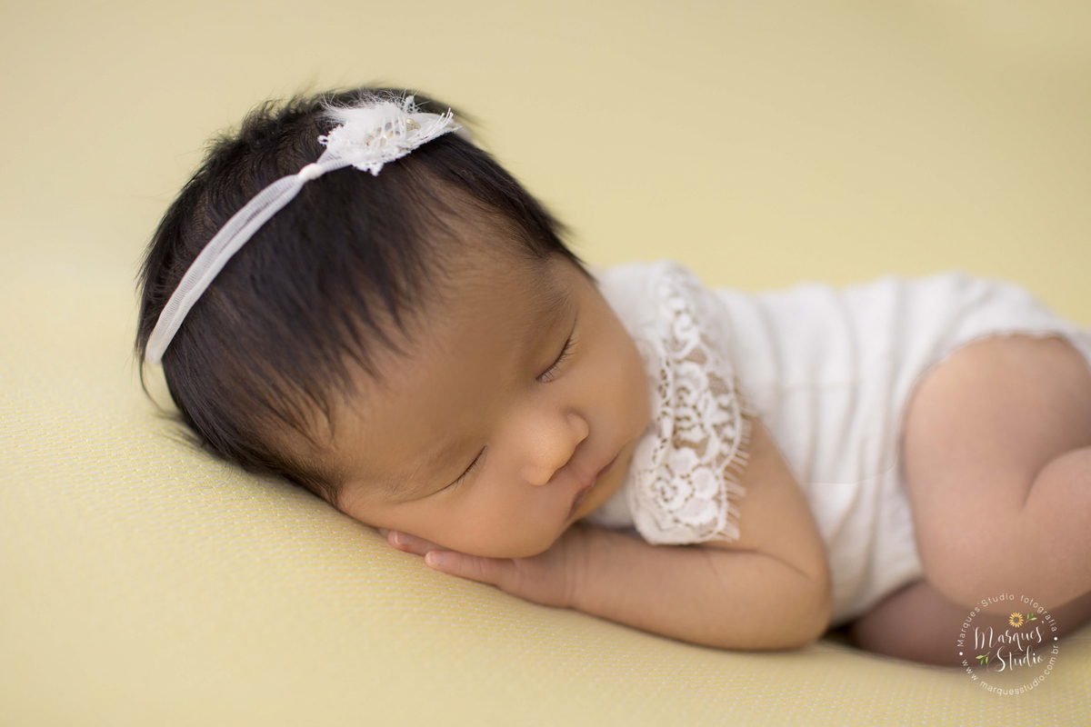 Newborn da Beatriz de 15 dias realizado em studio localizado na Av Paulista - São Paulo, SP. Na foto a bebê está deitada em um puff amarelo usando uma roupinha branca. Ela está com as mãozinhas embaixo do rosto em um close