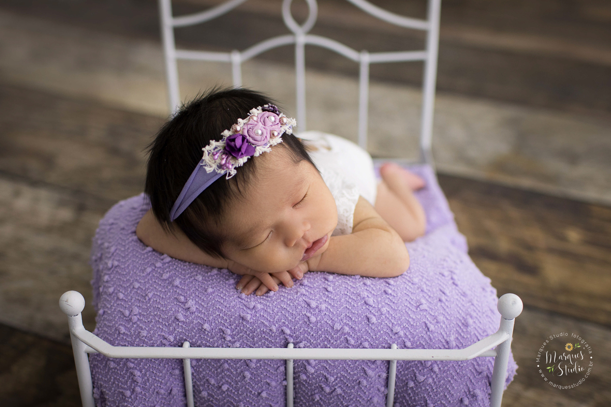 Foto realizada em ensaio newborn da Beatriz de 15 dias em studio localizado na Pompeia - São Paulo, SP. Na foto a bebê está deitada em uma caminha branca, como forro roxo, ela usa uma tiara roxa e está deitada de frente.