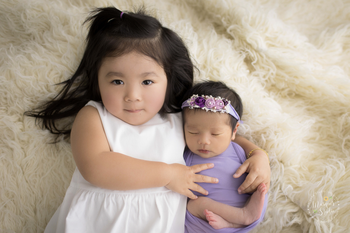 Ensaio realizado em studio localizado em Osasco - São Paulo, SP. Na foto a bebê está enrolada em um wrep roxo, deitada em um tapete branco sendo abraçada pela sua irmãzinha mais velha.