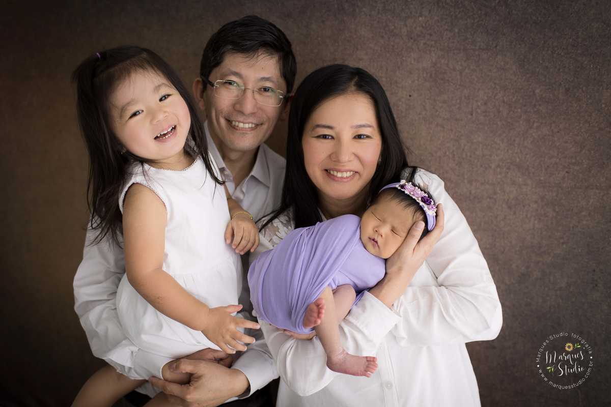 Ensaio realizado em studio localizado em Osasco - São Paulo, SP. Na foto a bebê está enrolada em um wrep roxo no colo da mamãe, enquanto o papai carrega sua irmã mais velha. Eles estão sorrindo e felizes.