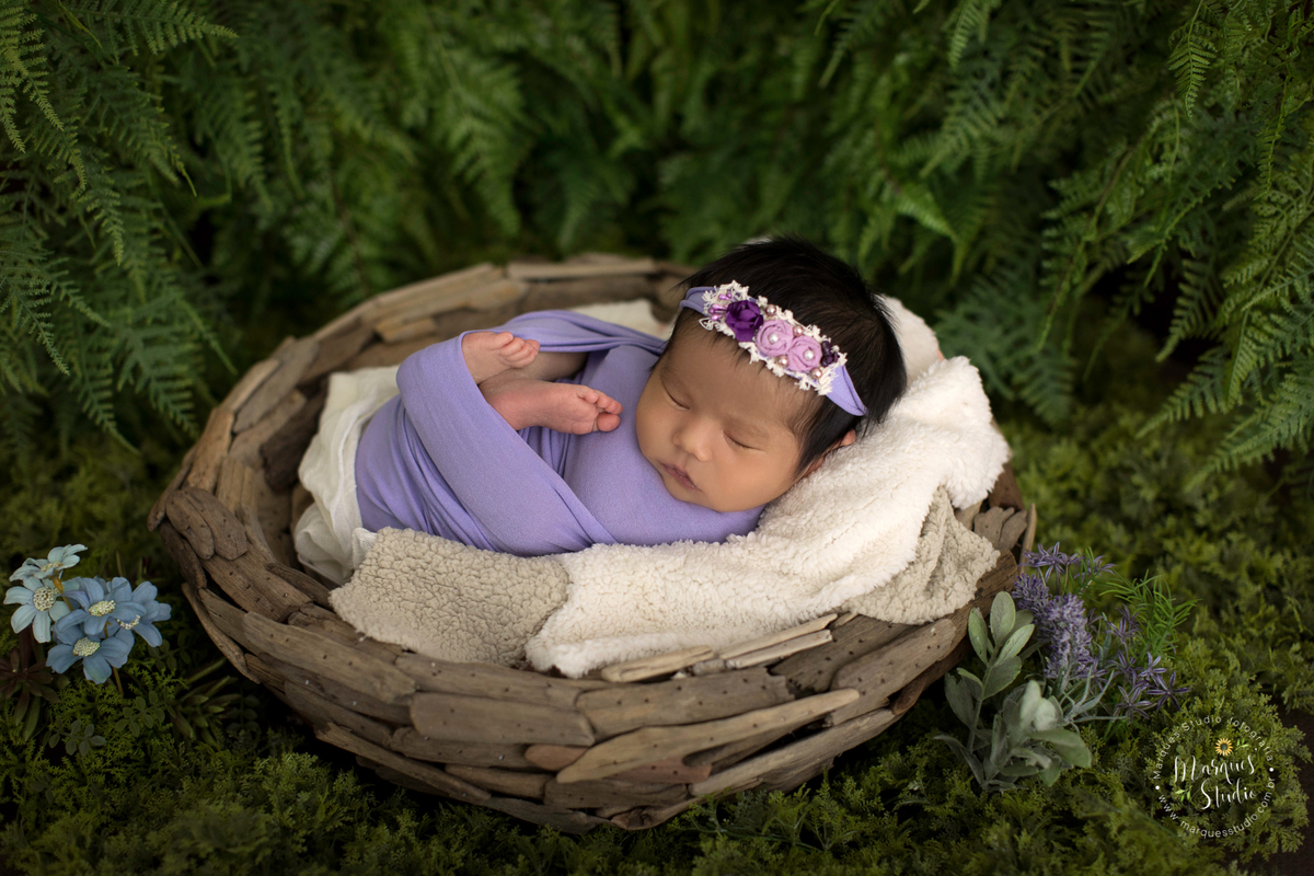 Foto realizada em ensaio newborn da Beatriz de 15 dias em studio localizado em Santo Amaro - São Paulo, SP. Na foto a bebê está deitada em um tronco de madeira, enrolada em um wrep roxo, o fundo fotográfico é um lindo jardim com flores.