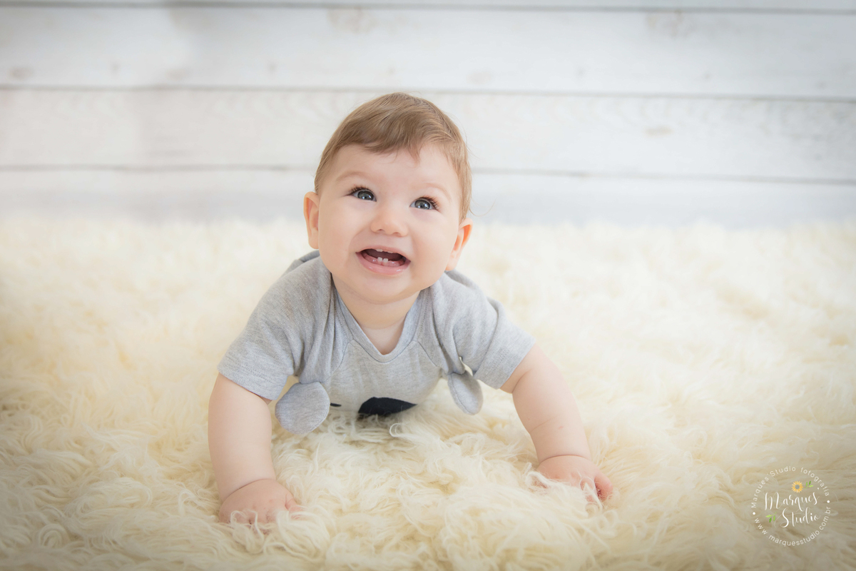 Foto feita em ensaio de bebê do Leonardo em studio localizado na Faria Lima - São Paulo, SP. Na foto o bebê está deitado de barriga pra baixo, em um tapete bege usando um blusa de bichinho, a foto foi tirada de cima para baixo e o bebê está sorrindo.