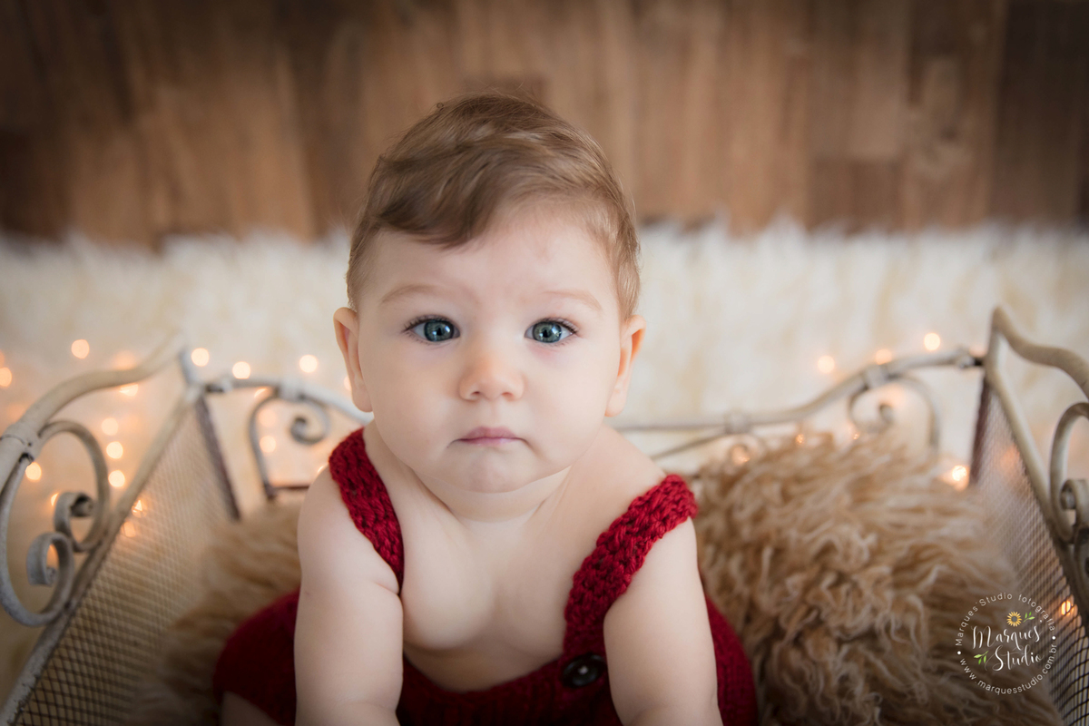 Foto feita em ensaio de bebê do Leonardo em studio localizado em Sumaré - São Paulo, SP. Na foto o bebê está sentado em um berço bege, usando um lindo macacão vermelho de crochê, a foto é um lindo close no rosto do bebê