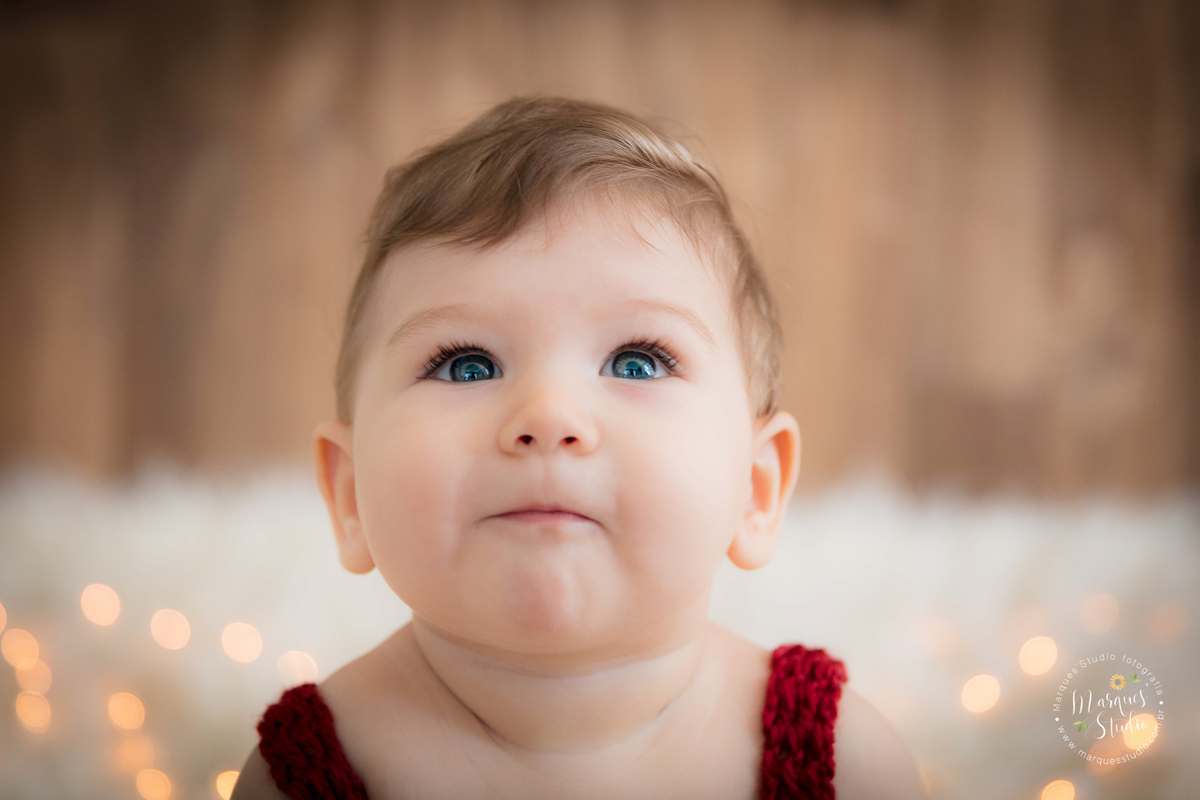 Foto feita em ensaio de bebê do Leonardo em studio localizado em Osasco - São Paulo, SP. Na foto o bebê está sentado em um berço bege, usando um lindo macacão vermelho de crochê, a foto é um lindo close no rosto do bebê que está sorrindo envergonhao