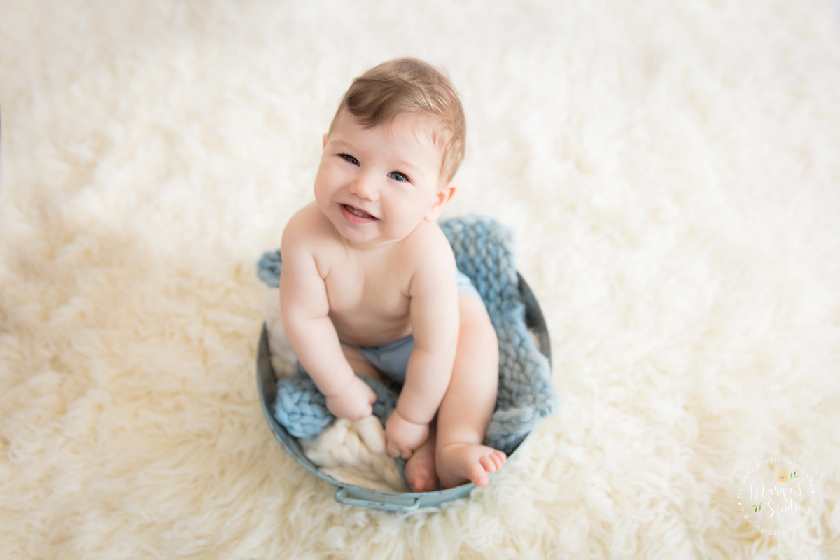 Foto feita em ensaio de bebê do Leonardo em studio localizado no Jardins - São Paulo, SP. Na foto o bebê está sentado em um balde azul com forro azul escuro, ele está pedalinho,  a foto foi tirada de cima para baixo e o bebê está sorrindo.