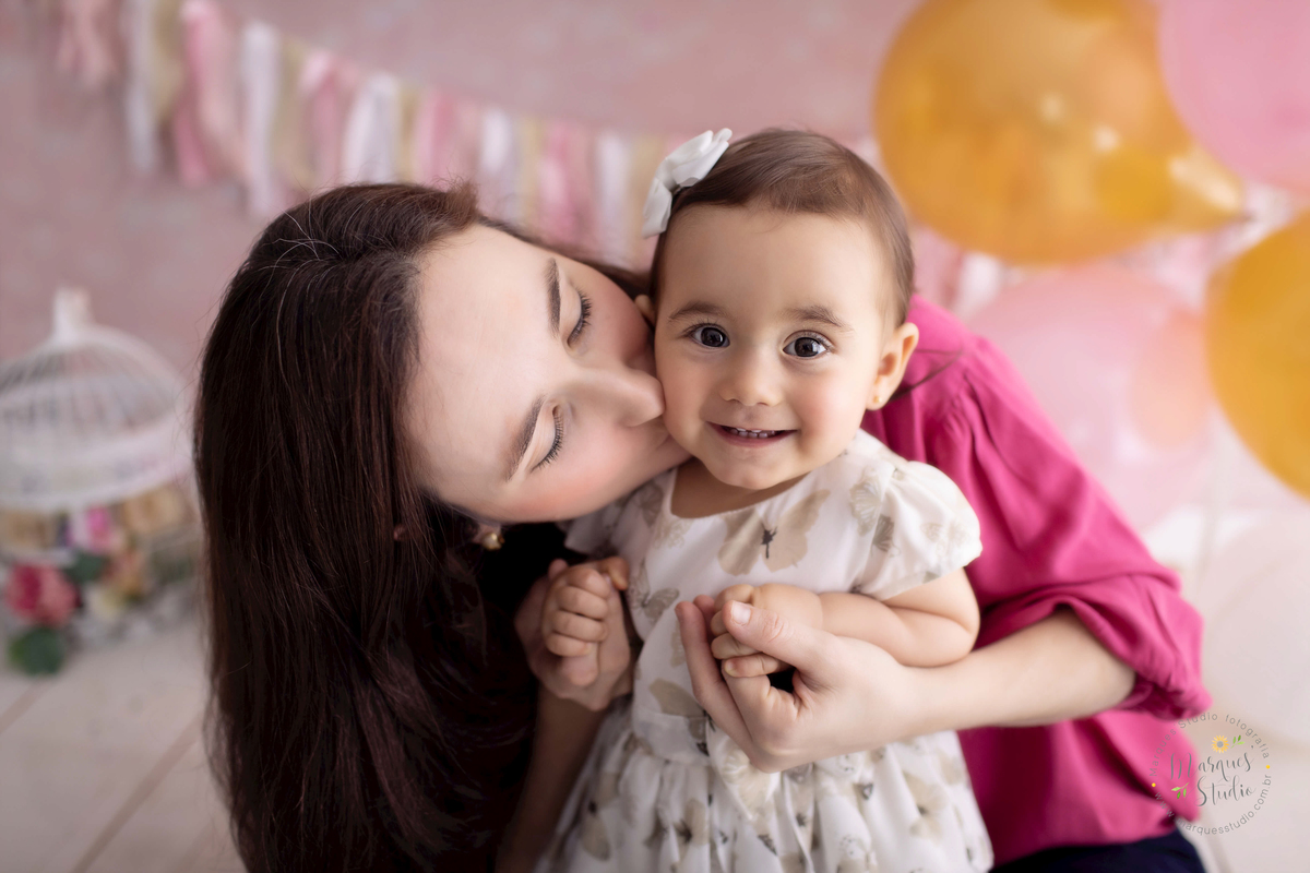Ensaio Smash the cake da Bia de um ano feito em studio fica localizado no Jardins - São Paulo, SP. Na foto temos a Bia sendo abraçada e beijada pela sua mamãe. ela está sorridente e feliz