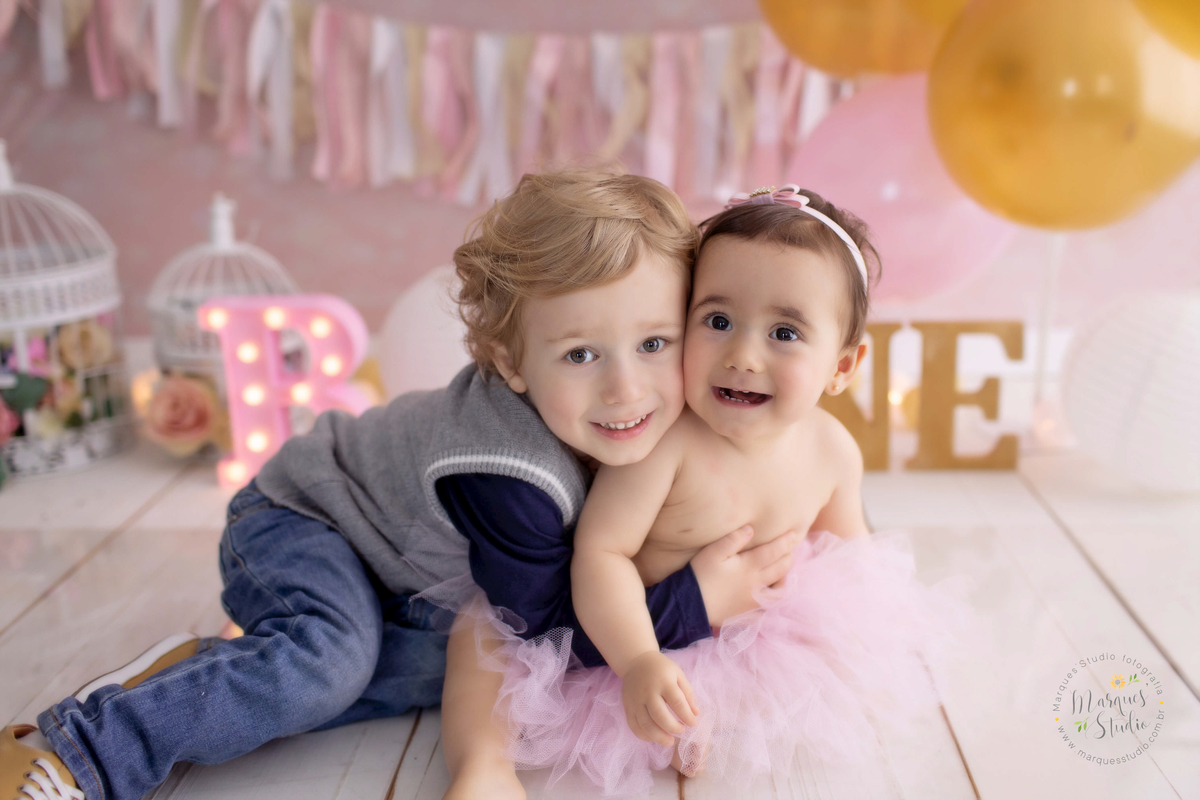 Fotografia feita no ensaio Smash the cake da Bia de um ano. Na foto ela está abraçada com seu irmão mais velho, os dois estão sorrindo. No fundo temos muitos balões coloridos e detalhes rosa. O studio fica localizado na Av Paulista - São Paulo, SP. 