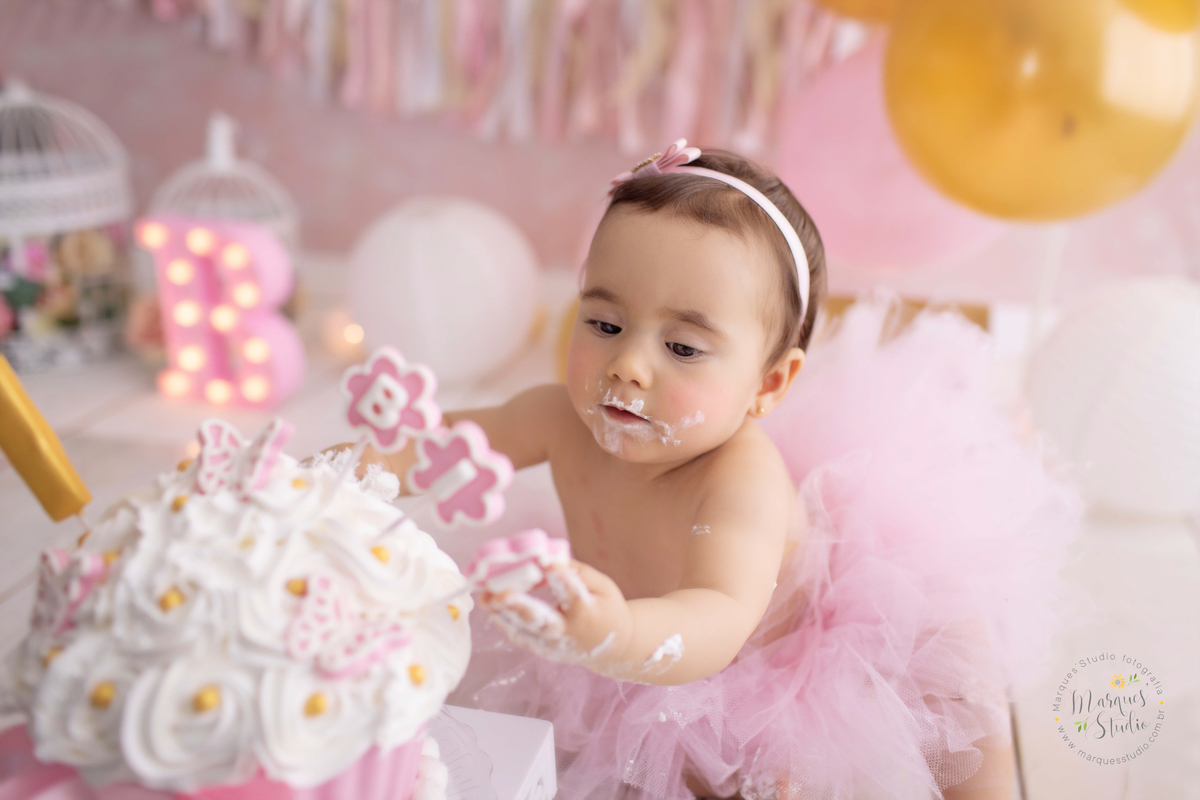 Foto feita em Ensaio Smash the cake da Bia de um ano. Na foto temos a Bia pegando no bolo com as mãos e o rosto bem sujos de glacê. O studio fica localizado em Osasco - São Paulo, SP. 