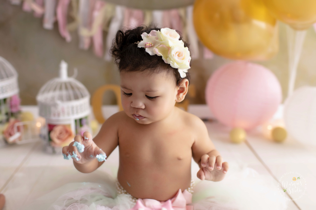 Ensaio feito em studio localizado na Av. Paulista - São Paulo, SP. Na foto temos a Marina no seu smash the cake. A bebê está usando uma tiara de flores no cabelo, está com as mãos sujas de bolo e tem uma lindo cenário rosa, dourada e branco atrás dela. 