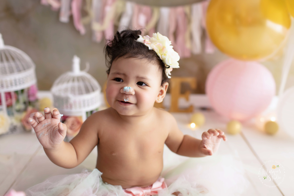 Fotografia feita em studio localizado na Vila Leopoldina  - São Paulo, SP. Na foto temos a Marina no seu smash the cake. A bebê está usando uma tiara de flores no cabelo, está com as mãos e o nariz sujo de bolo, sorrindo e se divertindo com seu bolo