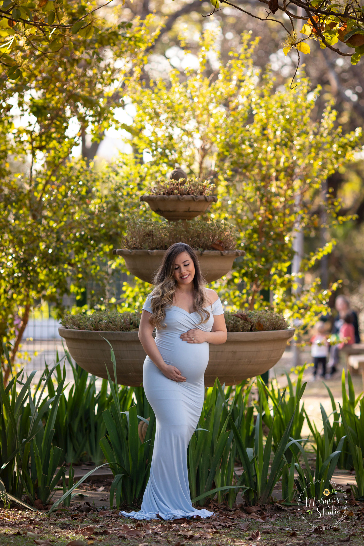 Foto feita para ensaio de gestante da Tatiana. Na foto a gestante está com as mãos na barriga e sorrindo, usando um lindo vestido azul. O ensaio foi realizado no parque em São Paulo, SP. 