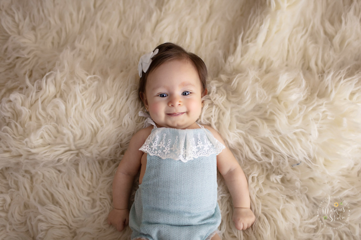 Ensaio realizado em studio localizado na Vila Leopoldina - São Paulo, SP. Na foto a Manuela de 3 anos está dando um lindo sorriso enquanto está deitada em um tapete branco, usando um macaquinho azul e um lacinho na cabeça