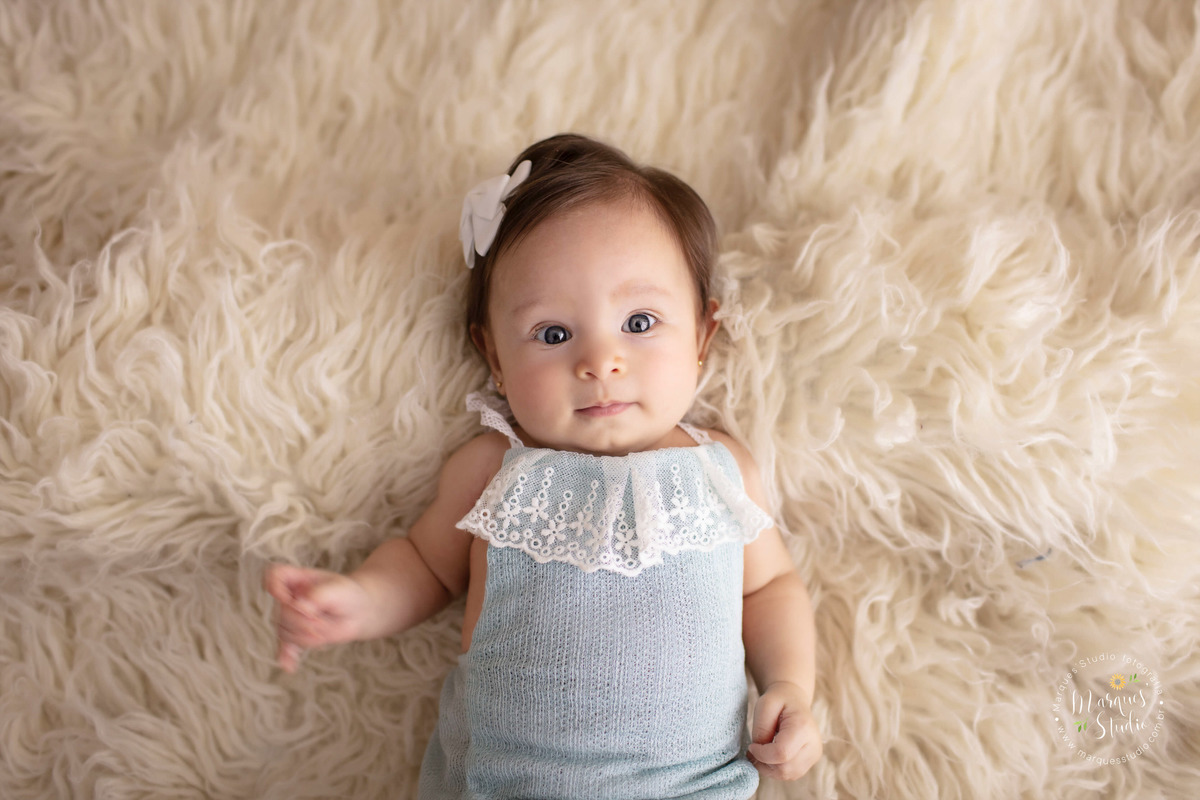 Ensaio realizado em studio localizado em Santo Amaro - São Paulo, SP. Na foto a Manuela de 3 anos está olhando fixamente  para câmera enquanto está deitada em um tapete branco, usando um macaquinho azul e um lacinho na cabeça