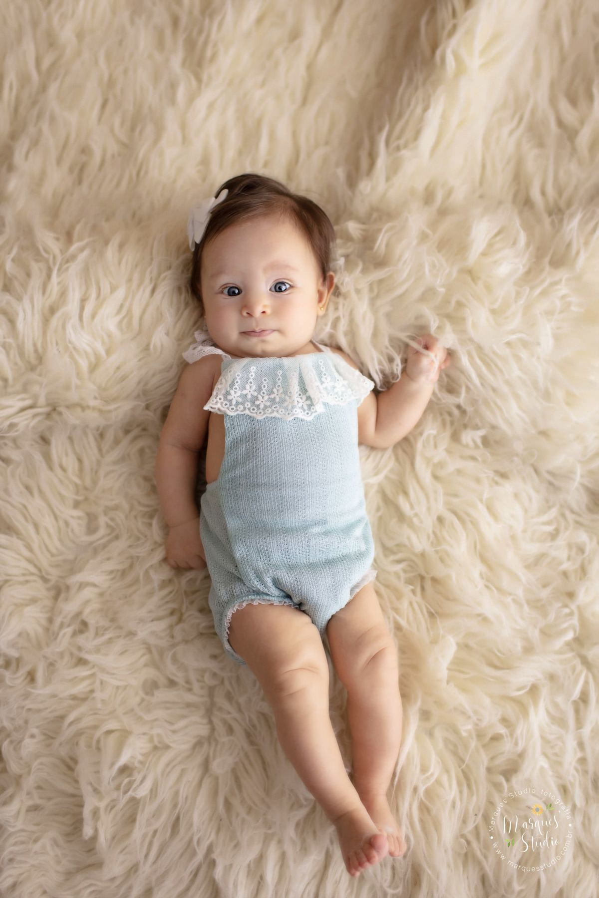 Ensaio feito em studio localizado na Ana Rosa - São Paulo, SP. Na foto a Manuela de 3 anos está deitada em um tapete branco, usando um macaquinho azul e um lacinho na cabeça. A foto mostra todo o corpo do bebê que olha fixamente para a câmera