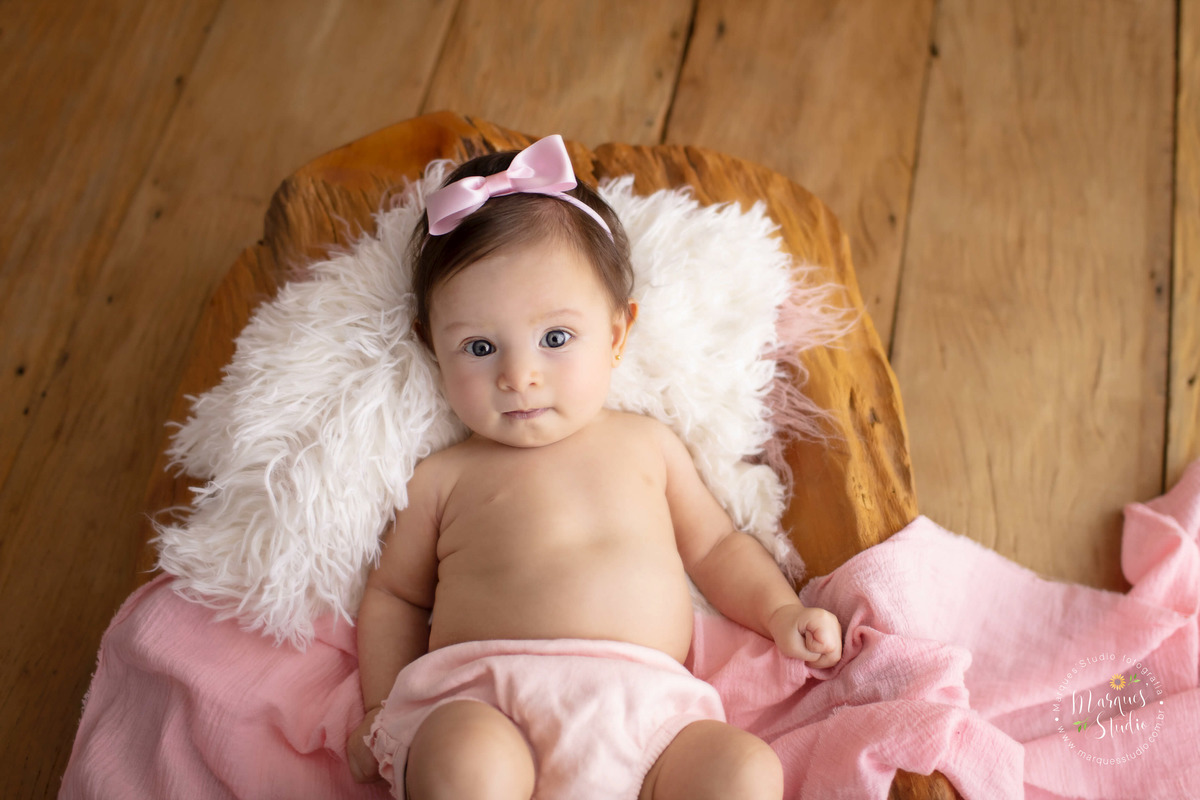 Foto feita no ensaios acompanhamento da Manuela de 3 anos. A foto é um close no rosto da bebê que está olhando fixamente para a câmera e está deitada em um tronco de madeira com um lenço rosa. Studio fica localizado em Sumaré - São Paulo, SP