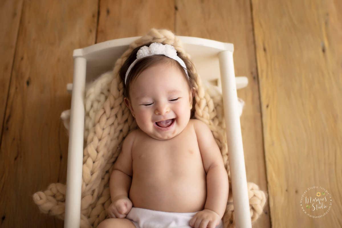 Foto feita no ensaios acompanhamento da Manuela de 3 anos. A foto é um close no rosto da bebê que está dando um lindo gargalhada de olhos fechados e está deitada em um berço branco. Studio fica localizado no Bela Vista - São Paulo, SP