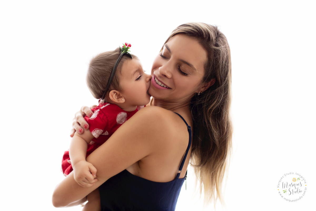 Foto da família da  Giovanna de 7 meses, feito em seu ensaio de acompanhamento em studio localizado no Bela Vista - São Paulo, SP. Na foto a Giovanna está sentada no colo da mamãe dando um beijão na bochecha dela, que está sorrindo e feliz 