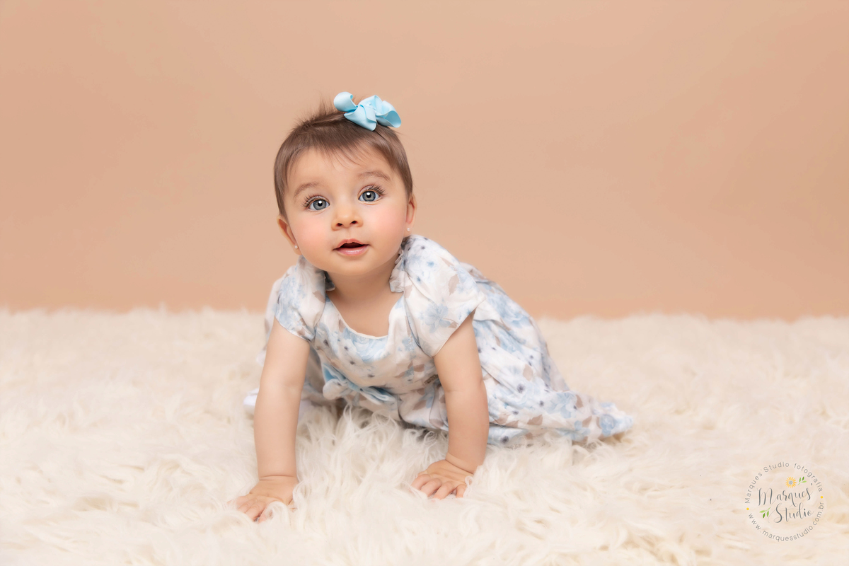 Foto da Giovanna de 7 meses, feito em studio localizado na Ana Rosa - São Paulo, SP. Na foto a Giovanna está engatinhando em um tapete branco, usando um vestido azul florido