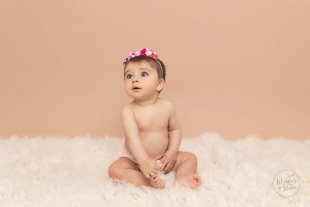 Foto da Giovanna de 7 meses, feito em studio localizado no Jardins - São Paulo, SP. Na foto a Giovanna está sentada em um tapete branco, usando uma calcinha rosa e uma flor no cabelo 