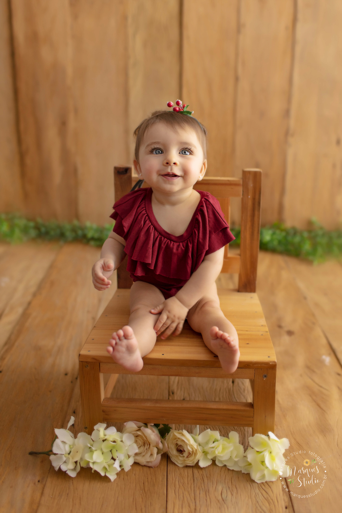 Foto da Giovanna de 7 meses, feito em studio localizado na Vila Prudente - São Paulo, SP. Na foto a Giovanna está sentada em uma cadeira de madeira, usando um macacão vermelho e sorrindo sapeca. 