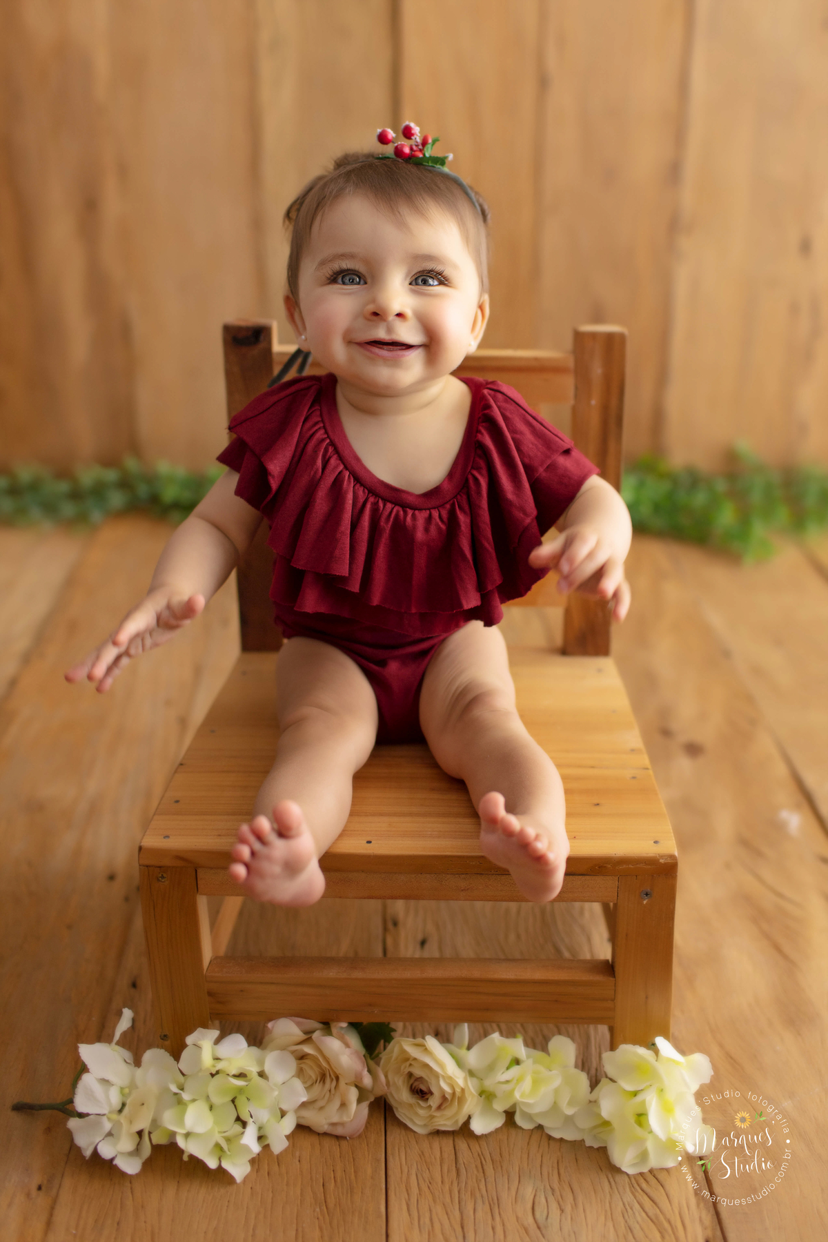 Foto da Giovanna de 7 meses, feito em studio localizado em Sumaré  - São Paulo, SP. Na foto a Giovanna está sentada em uma cadeira de madeira, usando um macacão vermelho e sorrindo sapeca. 