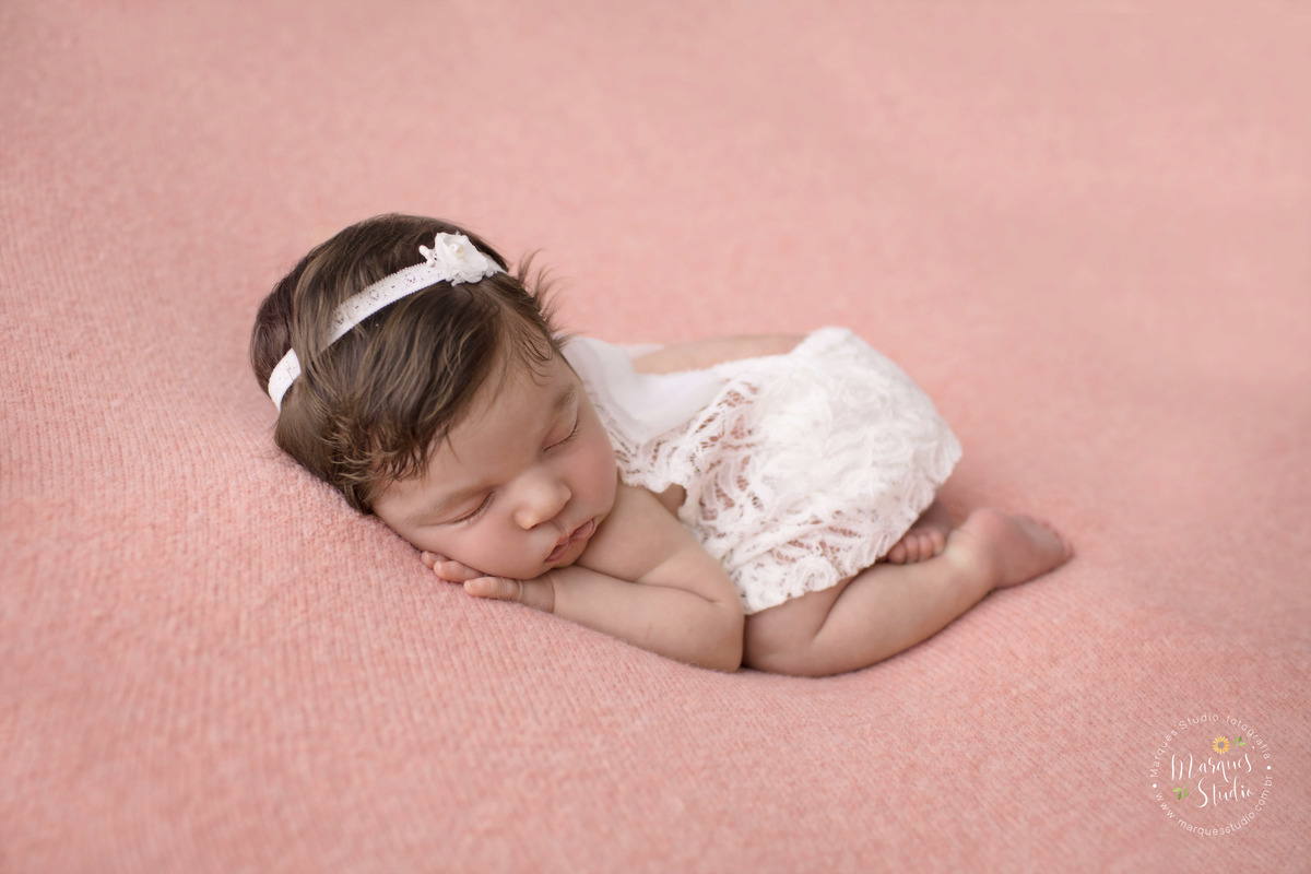 Newborn da Isabel de 16 dias realizado em studio localizado na Vila Madalena - São Paulo, SP. Na foto a bebê está deitada em um puff rosa com as mãos em baixo do rosto, usando um vestido branco e uma tiara no cabelo