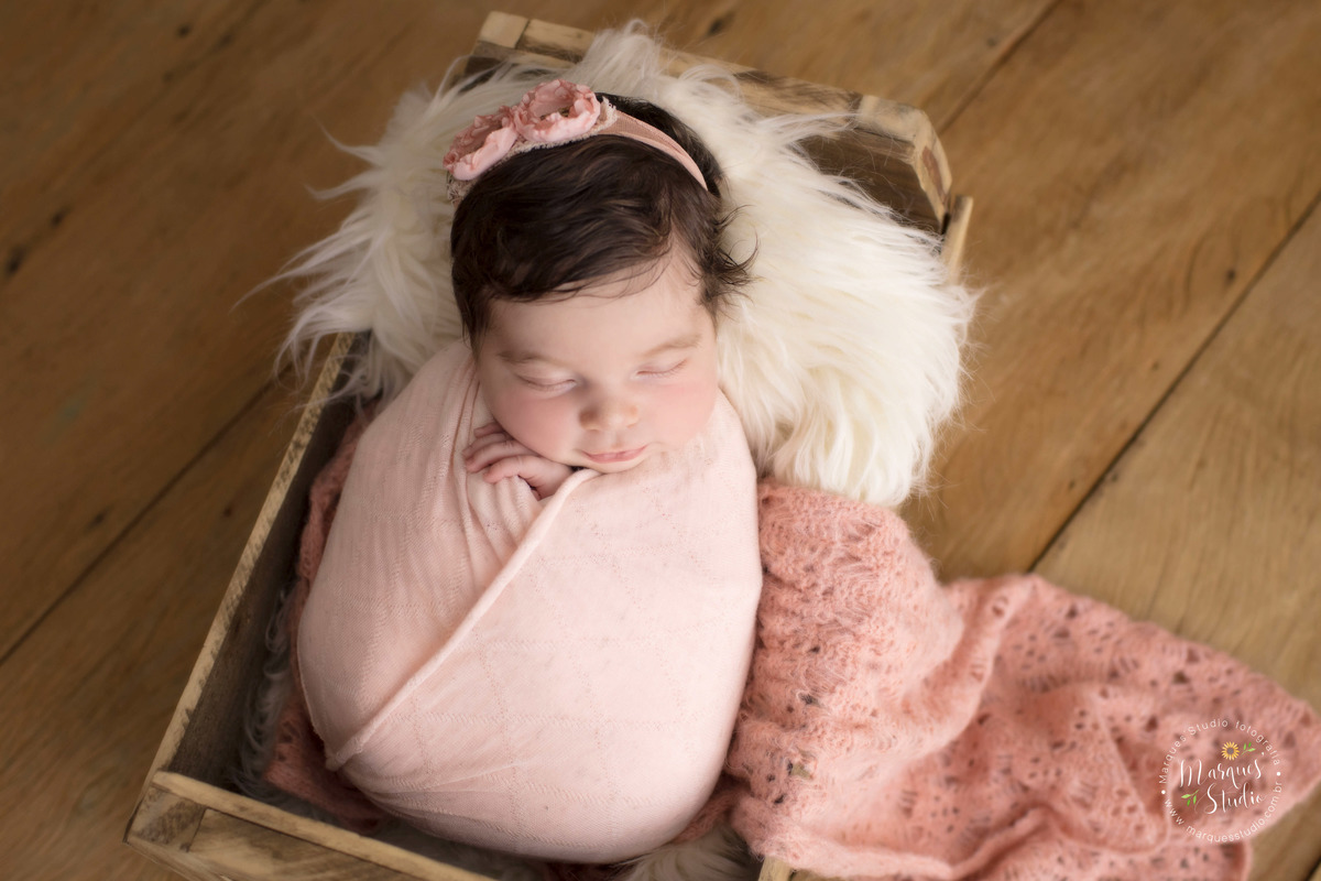 Foto feita para ensaio newborn em studio localizado em Santo Amaro - São Paulo, SP. Na foto a bebê está enrolada em um wrep rosa e está dentro de um baú de madeira com pelos brancos. 