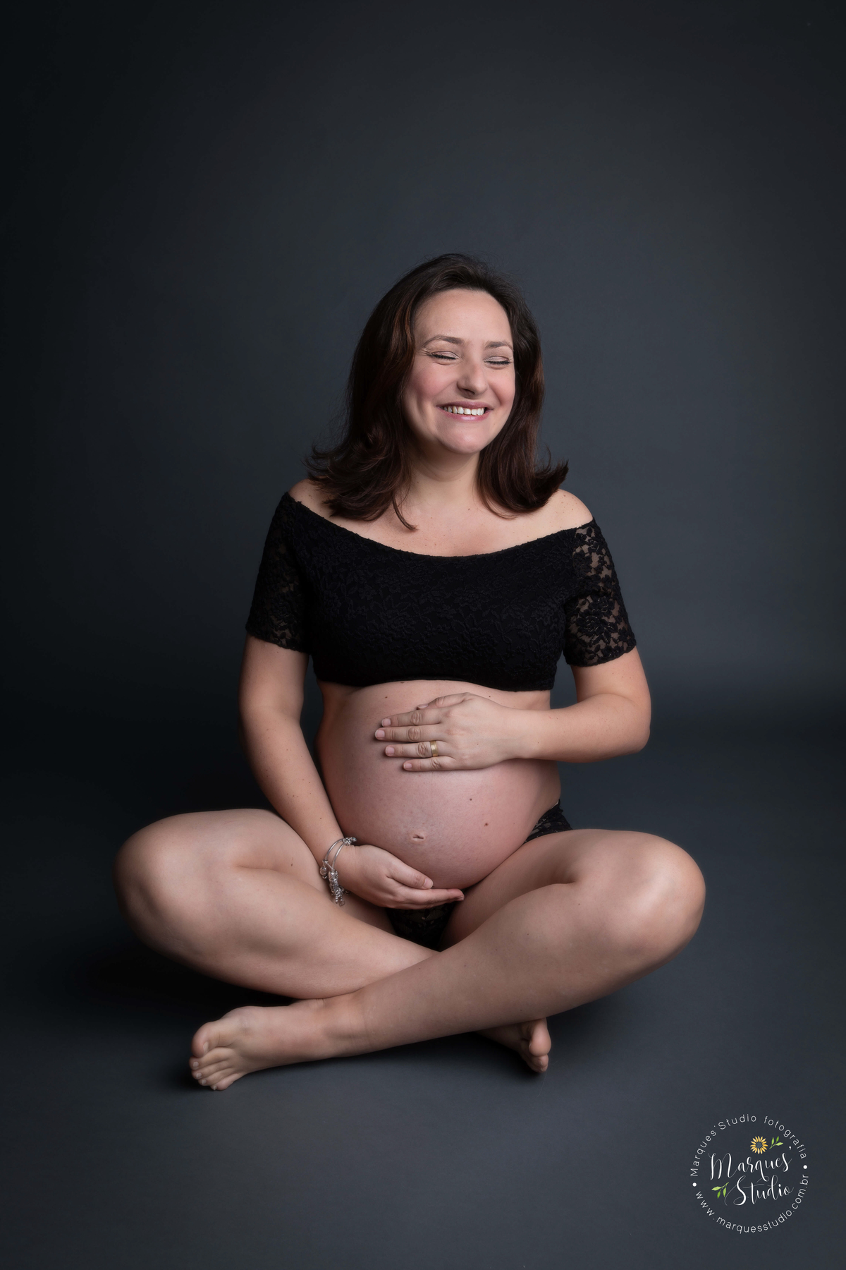 Foto feita em studio localizado na Zona Sul de São Paulo, SP. Na foto a mamãe está usando uma langeri preta, sentada em um fundo preto, ela está com as mãos na barriga e dando um lindo e largo sorriso