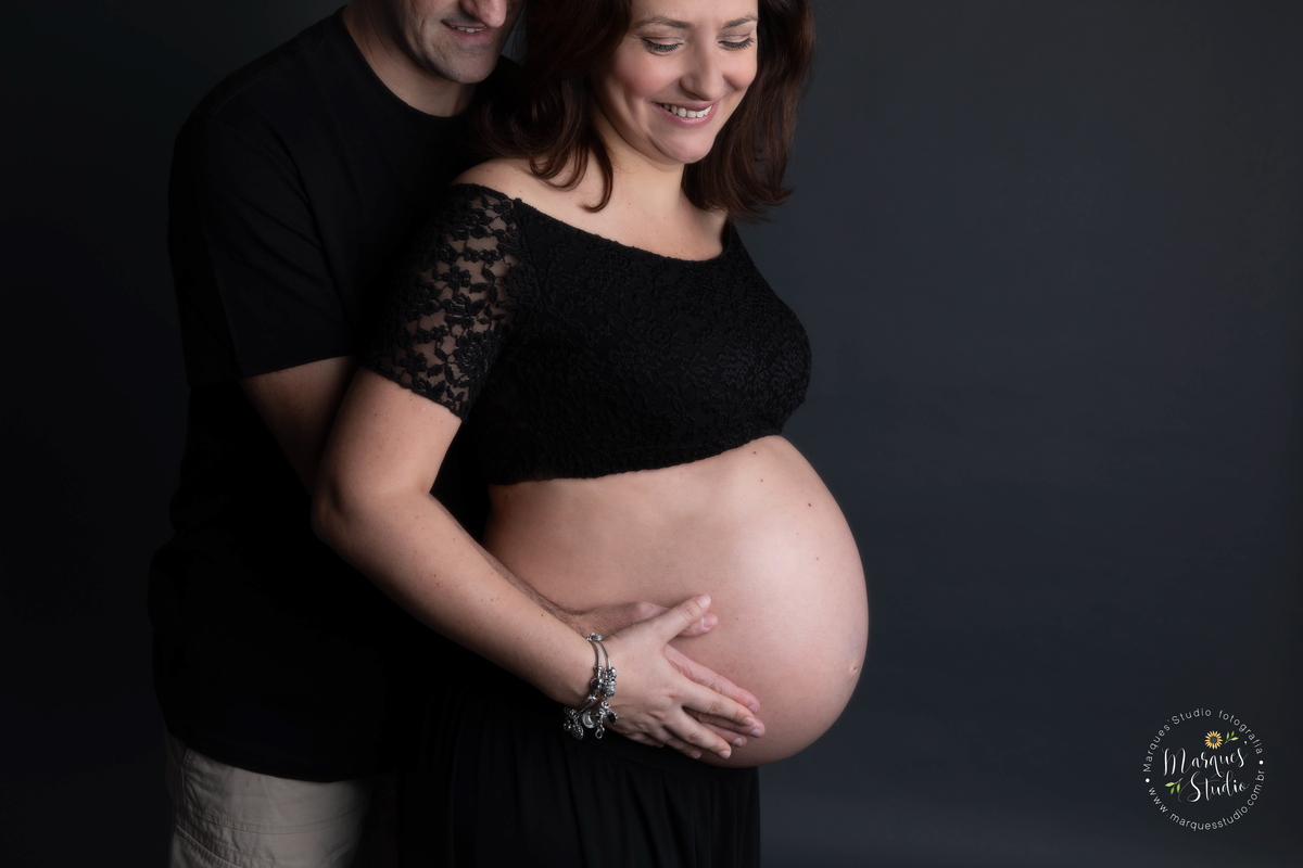 Ensaio fotografico realizado em studio localizado na Zona Oeste de São Paulo, SP. Na foto a mamãe está usando um conjunto de saia e top preto, enquanto seu marido a abraça por trás eles dão um lindo sorriso 