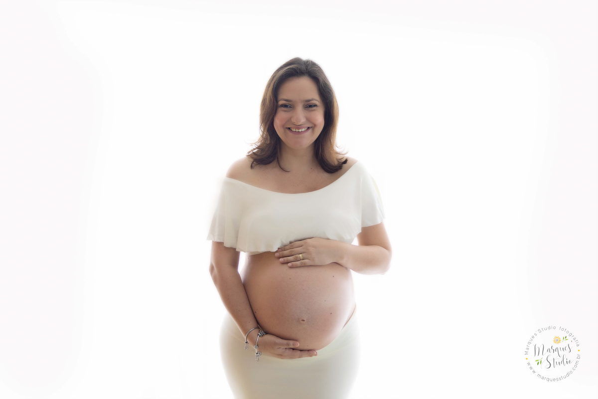 Ensaio fotografico realizado em studio localizado no Bela Vista - São Paulo, SP. Na foto a mamãe está usando um conjunto de saia e top branco, ela está com as mãos na barriga dando um lindo sorriso
