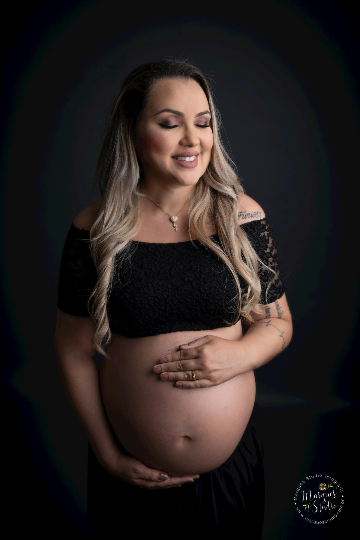 Foto feita para ensaio gestante da Micheli, em studio localizado em Osasco - São Paulo, SP. Na foto a mamãe está usando um conjunto de top e saia preta, com as mãos na barriga e dando um lindo sorriso para o seu bebê