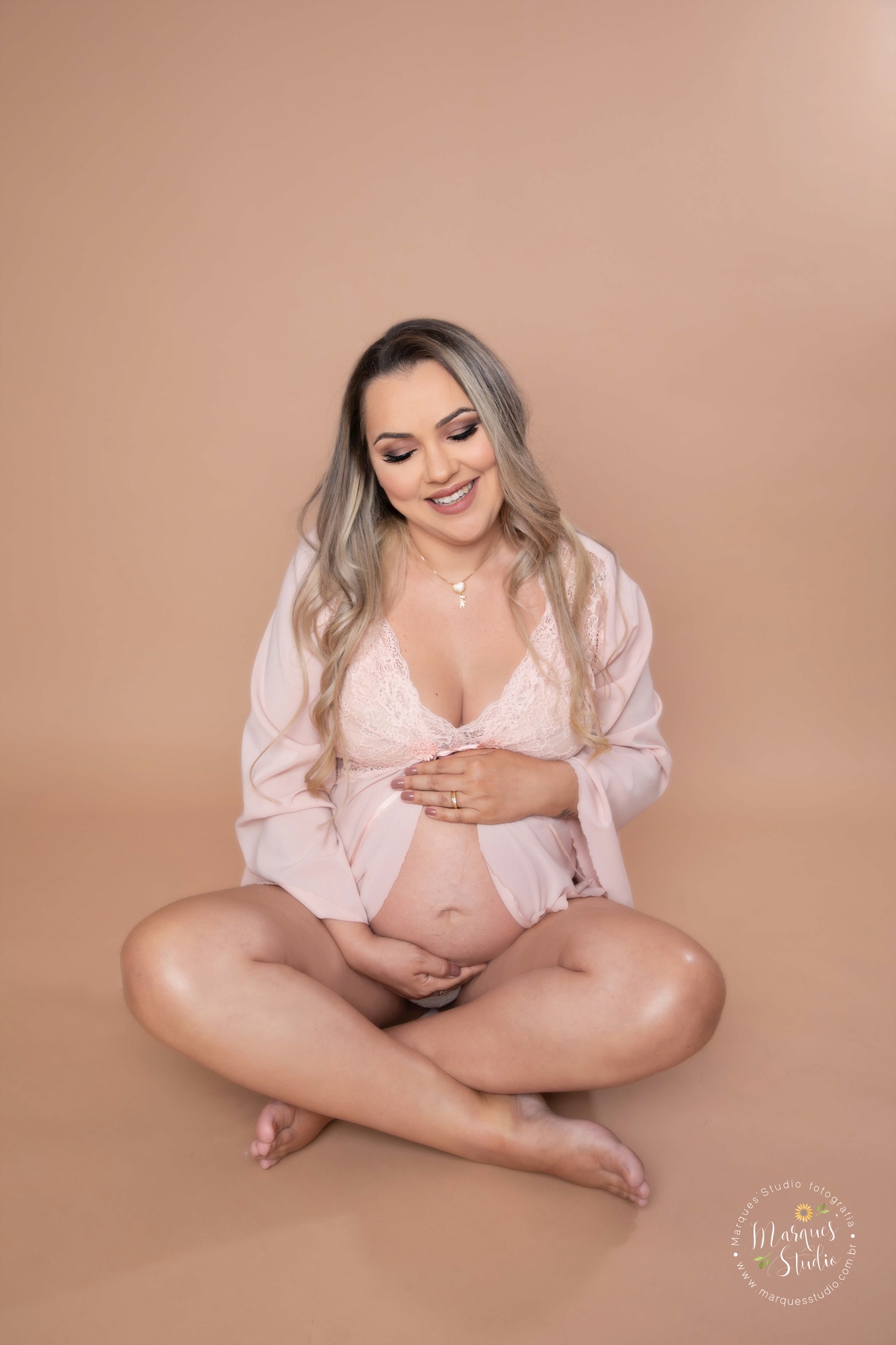 Foto feita para ensaio gestante da Micheli, em studio localizado no Jardins - São Paulo, SP. Na foto a mamãe está usando uma camisola rosa, com as mãos na barriga e dando um lindo sorriso para o seu bebê, ela está sentada no chão 