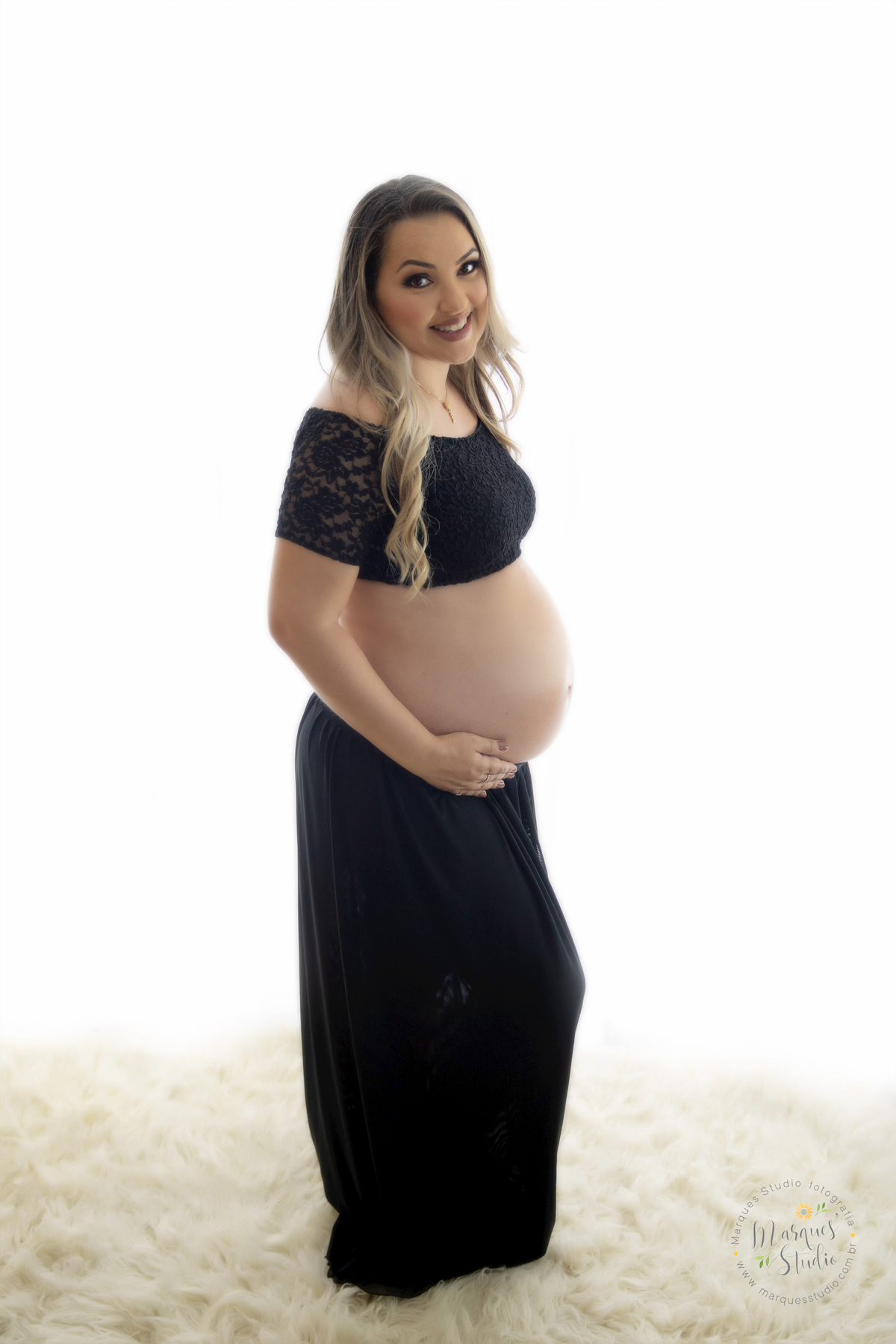 Foto feita para ensaio gestante da Micheli, em studio localizado em Sumaré - São Paulo, SP. Na foto a mamãe está usando um conjunto de top e saia preta, com as mãos na barriga e dando um lindo sorriso para o seu bebê