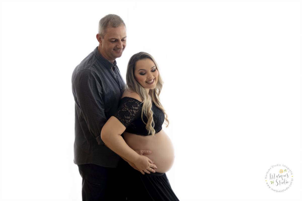 Ensaio de gestante realizado em studio localizado em Santo Amaro - São Paulo, SP. Na foto a mamãe está segurando a barriga enquanto o papai abraça ela por trás. Os dois estão dando um lindo sorriso