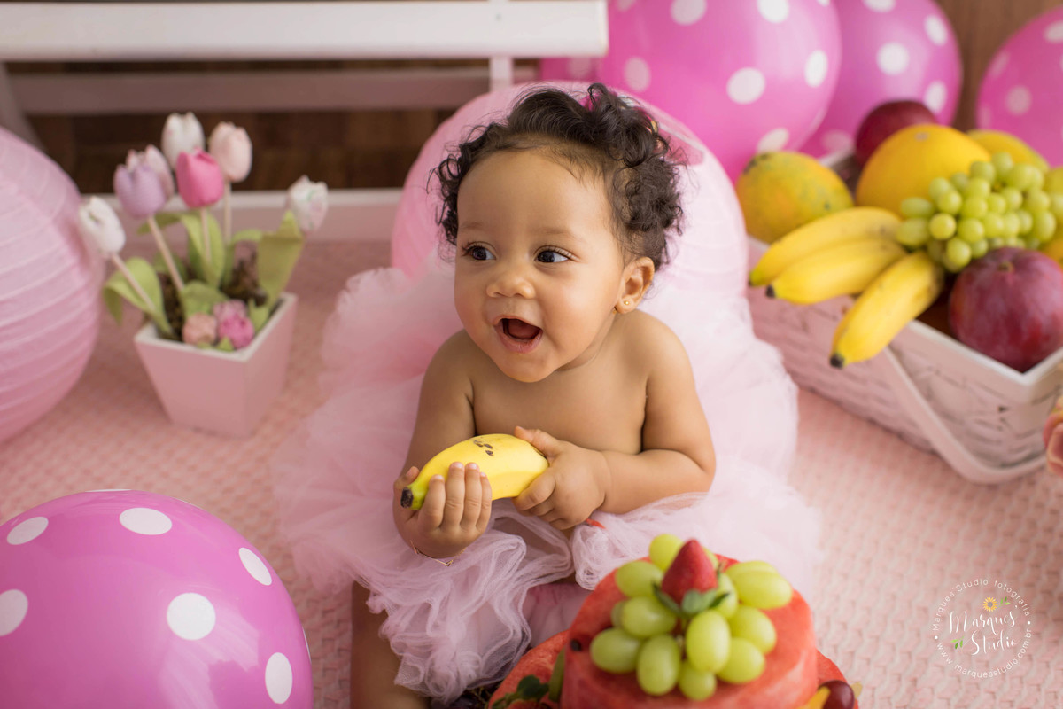 Ensaio smash the fruit feito em studio que encontra-se localizado em Sumaré - São Paulo, SP. Na foto a bebê Tayala está rindo e segurando uma banana bem amarela, em frente ao seu lindo bolo de frutas. 