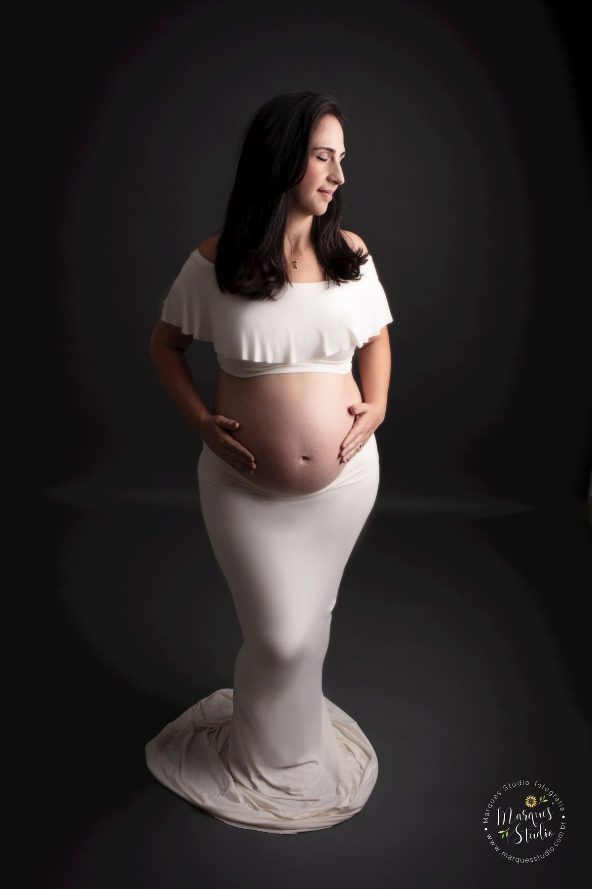 Ensaio de gestante da Renata realizado em studio localizado em Osasco - São Paulo, SP. Na foto a futura mamãe está com as mãos na barriga e olhando para a luz, em uma foto artistica. Ela usa um top e saia branco