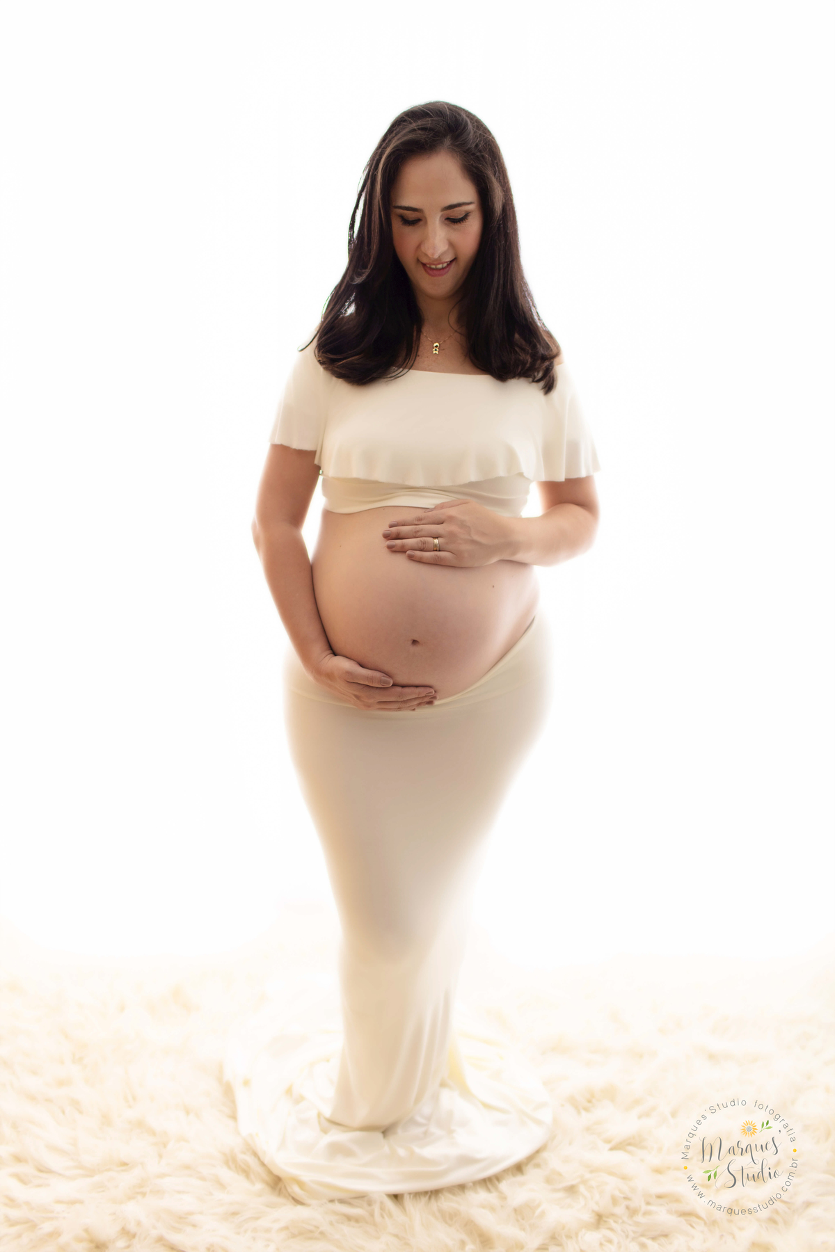 Ensaio de gestante da Renata realizado em studio localizado em Sumaré - São Paulo, SP. Na foto a futura mamãe está com as mãos na barriga e olhando para o seu bebê. Ela usa um top e saia branco