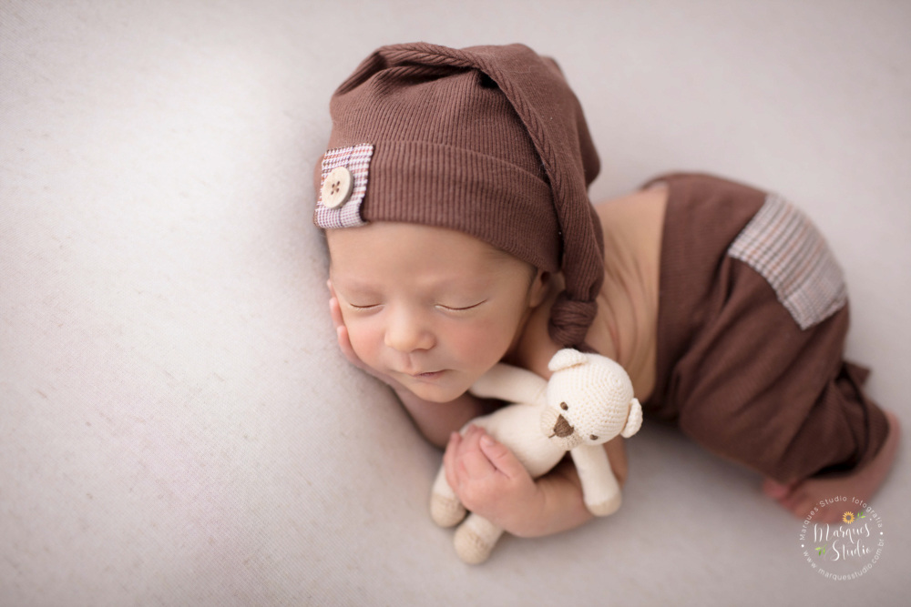 Ensaio feito em studio localizado na Pompeia - São Paulo, SP. Na foto o bebê está deitado de bundinha pra cima, com uma roupinha marrom, está deitado em um puff bege e abraçado um um risinho de pelúcia 