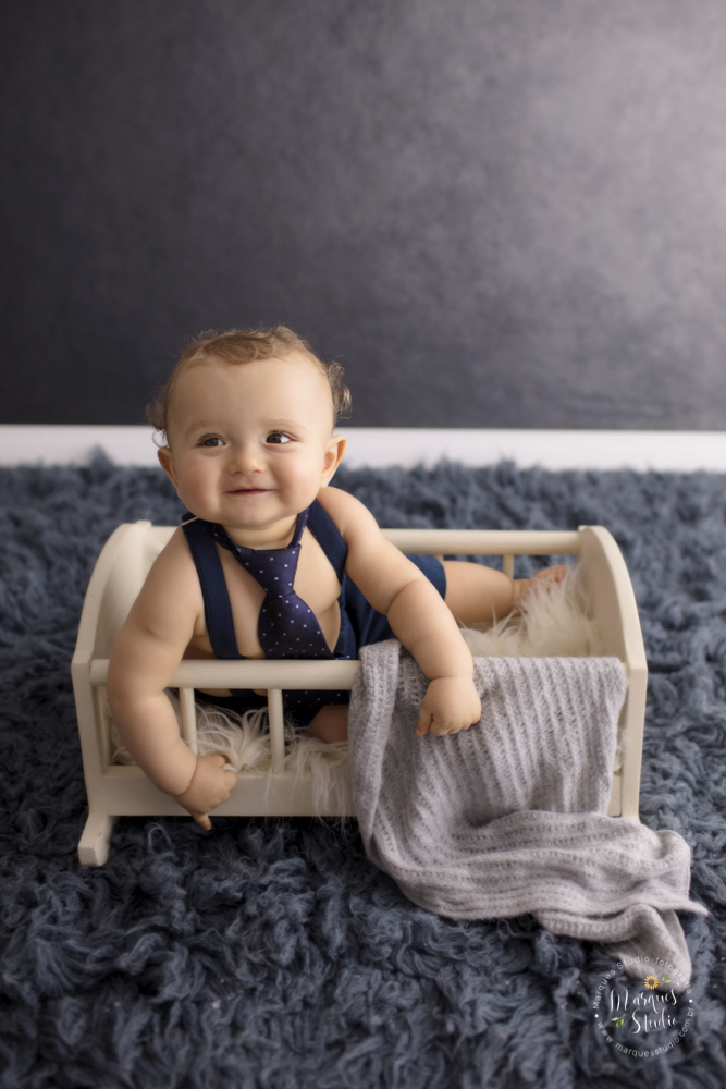 Foto do Enrico tirada em seu ensaio  de 1 ano em studio localizado Zona Sul de São Paulo, SP. Na foto o bebê está sentado em um bercinho branco ele está dando um lindo sorrio, O fundo fotográfico é uma parede azul e um tapete também azul