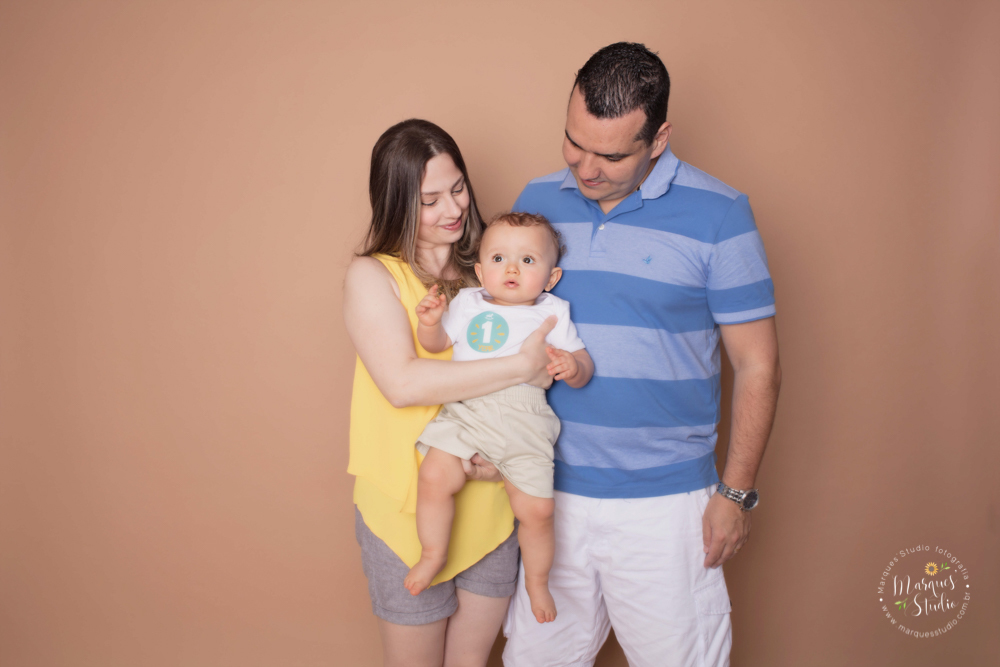 Foto da família do Enrico tirada em seu ensaio  de 1 ano em studio localizado na Zona Oeste de São Paulo, SP. Na foto o bebê está no colo da mamãe enquanto ela e o papai olham pra ele com amor e ternura. 