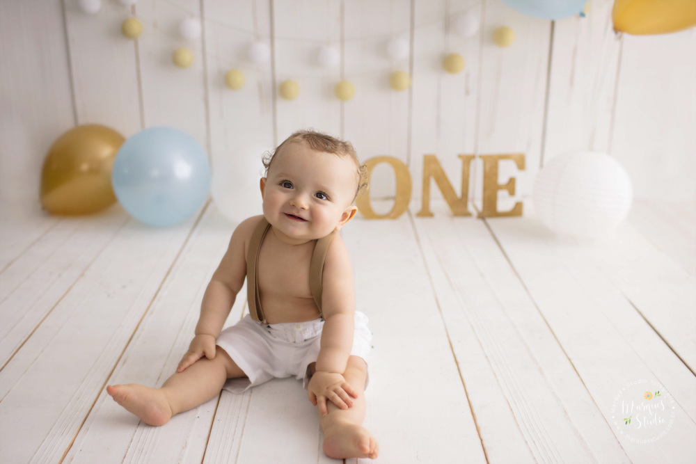 Ensaio de bebê do Enrico de 1 ano em studio localizado em Sumaré - São Paulo, SP. Na foto o bebê está sentado no chão dando um lindo sorriso, o fundo fotográfico é de madeira com balões e o nome 