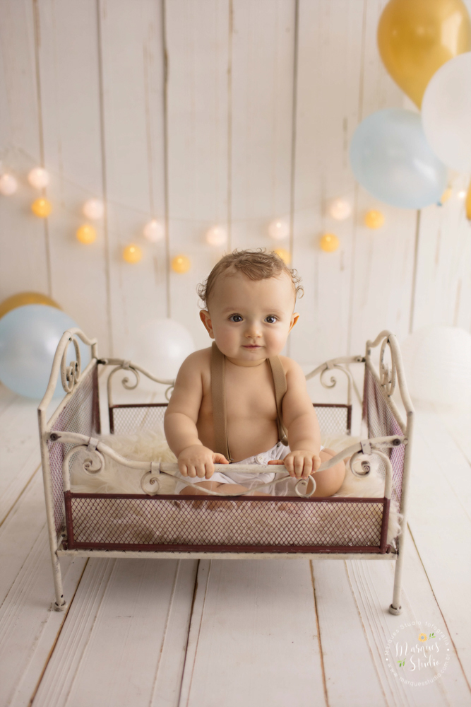 Ensaio de bebê do Enrico de 1 ano em studio localizado na Av. Paulista - São Paulo, SP. Na foto o bebê está sentado em um bercinho e dando um lindo sorriso, o fundo fotográfico é de madeira com balões e o nome 