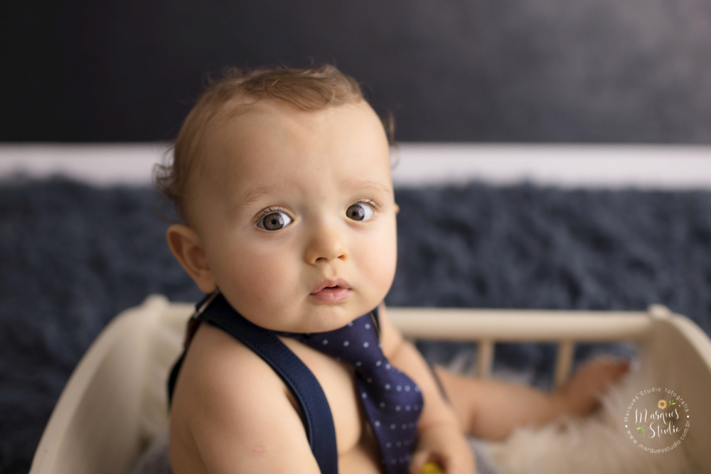 Foto do Enrico tirada em seu ensaio  de 1 ano em studio localizado em Santo Amaro - São Paulo, SP. Na foto o bebê está sentado em um bercinho branco, a foto é um close no seu rostinho, O fundo fotográfico é uma parede azul e um tapete também azul