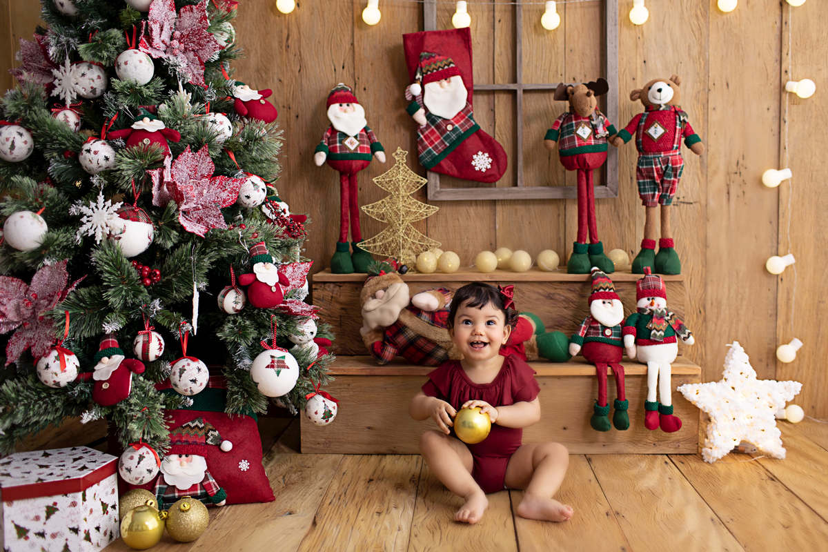 Ensaio de Natal da Laura de 1 ano em studio localizado em Santo Amaro - São Paulo, SP. Na foto a bebê está sentada no meio do cenário de natal, ela está usando um body de cor vinho e segurando um a bolinha da árvore de natal