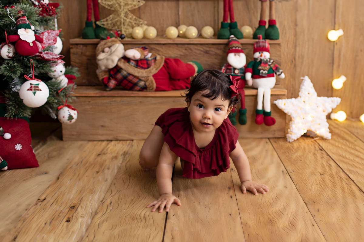 Fotografia feita no ensaio de Natal da Laura de 1 ano em studio localizado em Perdizes - São Paulo, SP. Na foto a bebê está engatinhando no meio do cenário de natal, ela está usando um body de cor vinho 